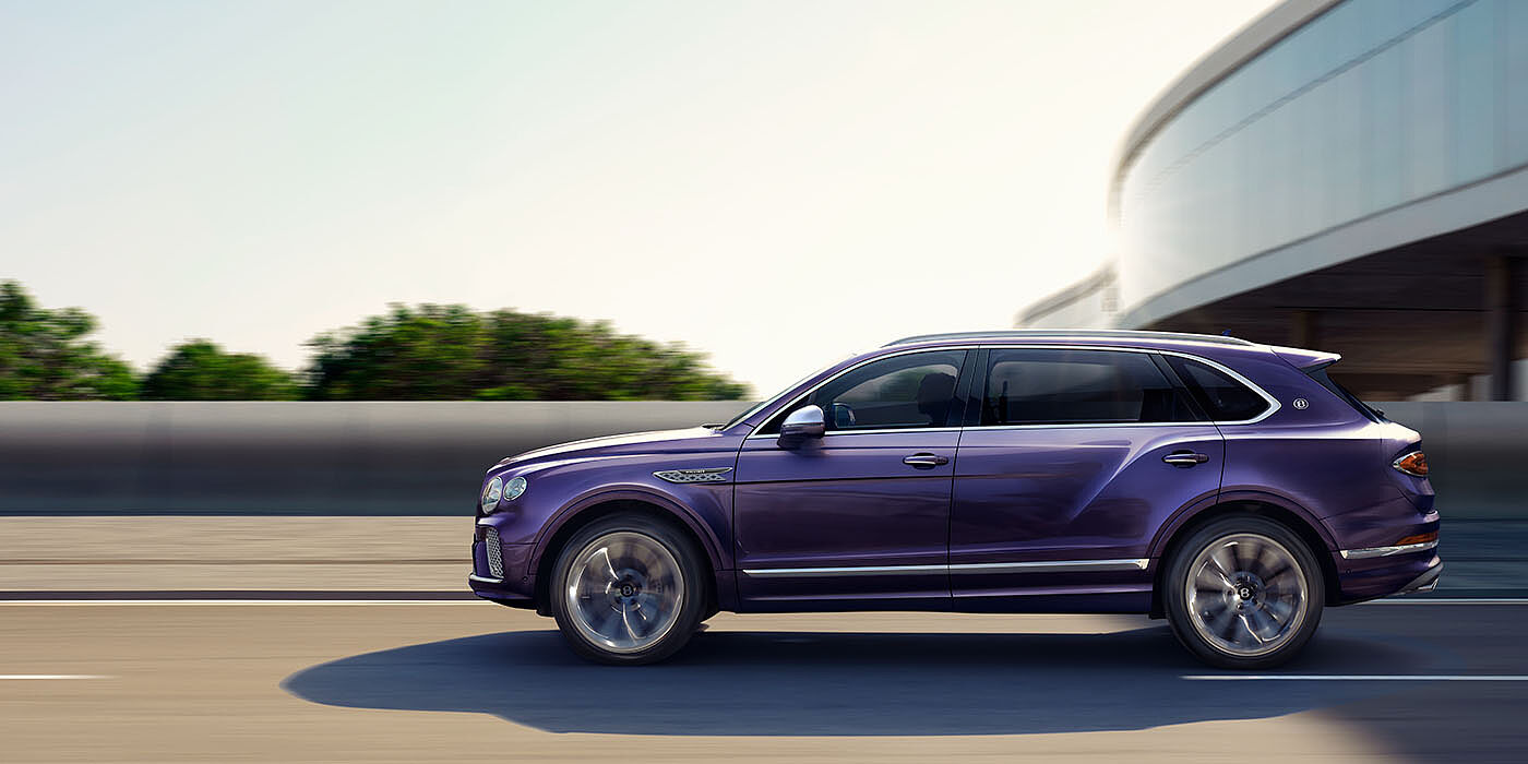 Bentley Bristol Bentley Bentayga Extended Wheelbase Mulliner luxury SUV profile in Tanzanite Purple paint, driving on an country road past trees.