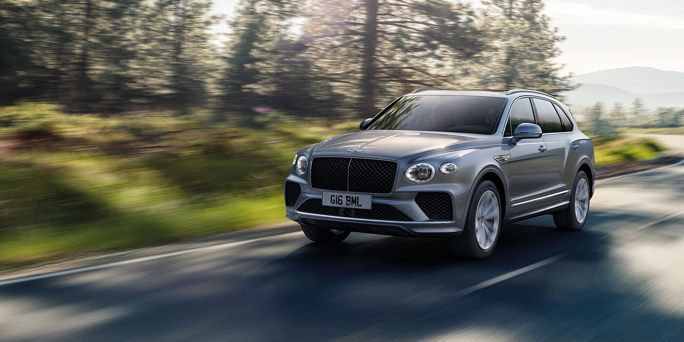Bentley Bristol Bentley Bentayga luxury SUV front three quarter in Moonbeam paint, driving on an country road past trees.